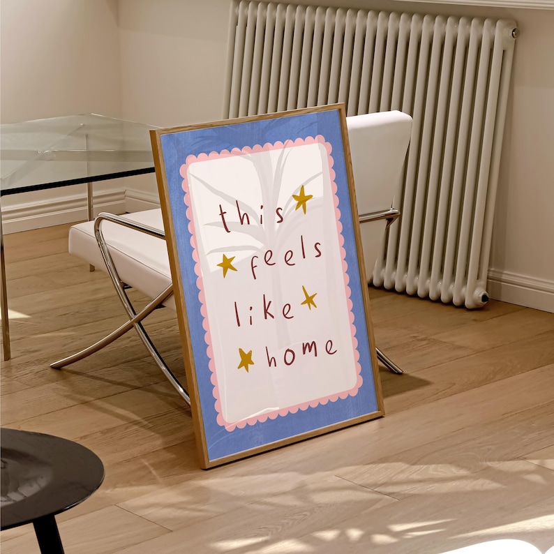 K&ouml;nnte beinhalten: Ein gerahmter Druck mit blauem Hintergrund und wei&szlig;em Rahmen. Der Druck zeigt den Text "this feels like home" mit Sternen um die W&ouml;rter.