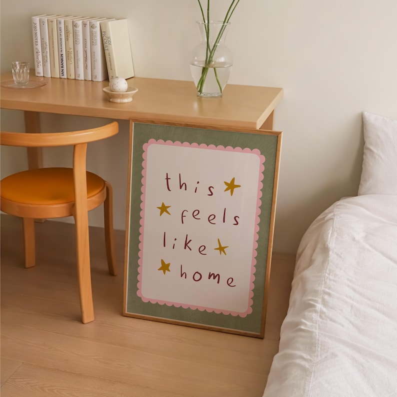 K&ouml;nnte beinhalten: Ein gerahmter Druck mit einem rosa und gr&uuml;nen Rahmen und dem Text "this feels like home" in braunen Buchstaben mit goldenen Sternen.