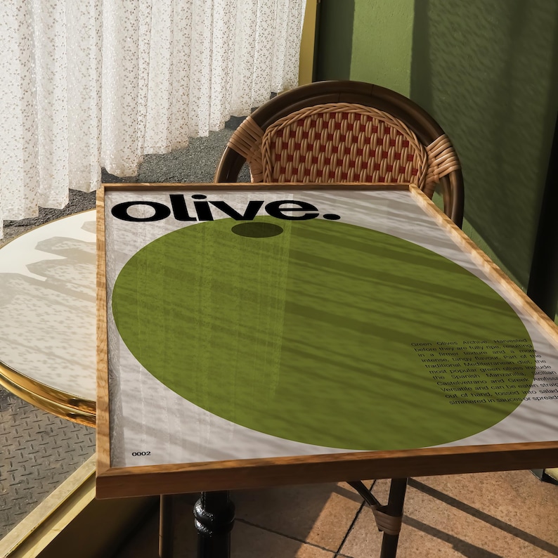 May include: A framed poster with the word "olive." in black text on a white background. A large green circle with a smaller black circle in the centre is behind the text. The poster is on a wooden table.