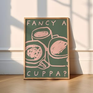 Könnte beinhalten: Gerahmter Kunstdruck mit grünem Hintergrund und rosa Strichzeichnungen von drei Kaffeetassen. Die Worte "FANCY A CUPPA?" sind in Grün oben und unten auf dem Bild gedruckt. Der Rahmen ist aus hellem Holz.
