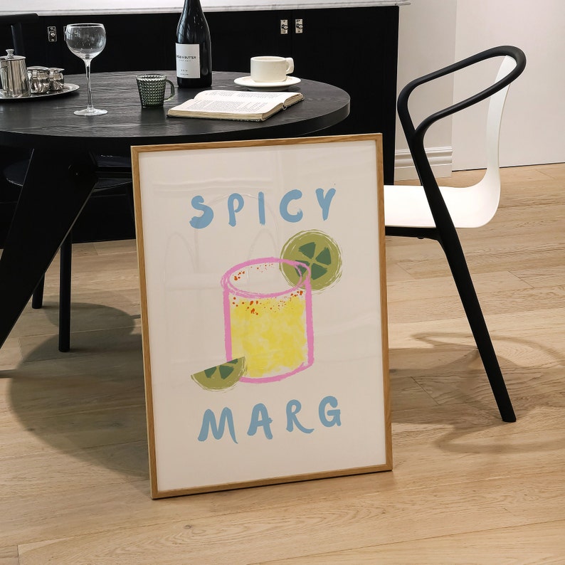 May include: A framed art print featuring a colorful illustration of a margarita cocktail. The artwork includes the words "SPICY MARG" in blue, with a yellow and pink cocktail design and lime garnish. The frame is a light brown color.