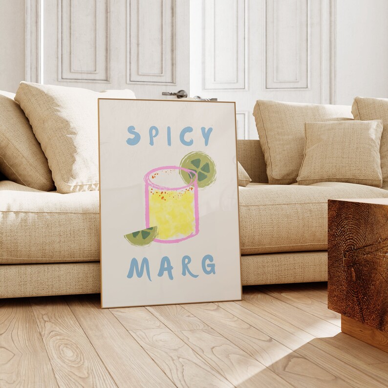 May include: A framed art print featuring a colorful illustration of a margarita cocktail. The words "SPICY MARG" are written in blue above and below the drink. The drink is yellow with a pink outline and a lime wedge.