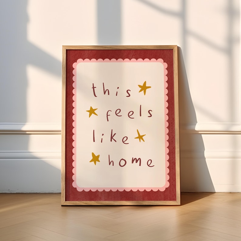 K&ouml;nnte beinhalten: Ein gerahmter Druck mit rotem Hintergrund und rosa Rand. Der Druck zeigt den Text "this feels like home" mit goldenen Sternen.