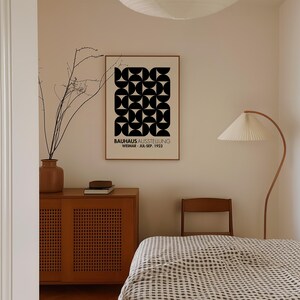 May include: A black and white geometric poster with the text "BAUHAUS AUSSTELLUNG WEIMAR - JUL-SEP. 1923" hanging on a beige wall in a room with a wooden cabinet, a chair, and a bed with a checkered bedspread.