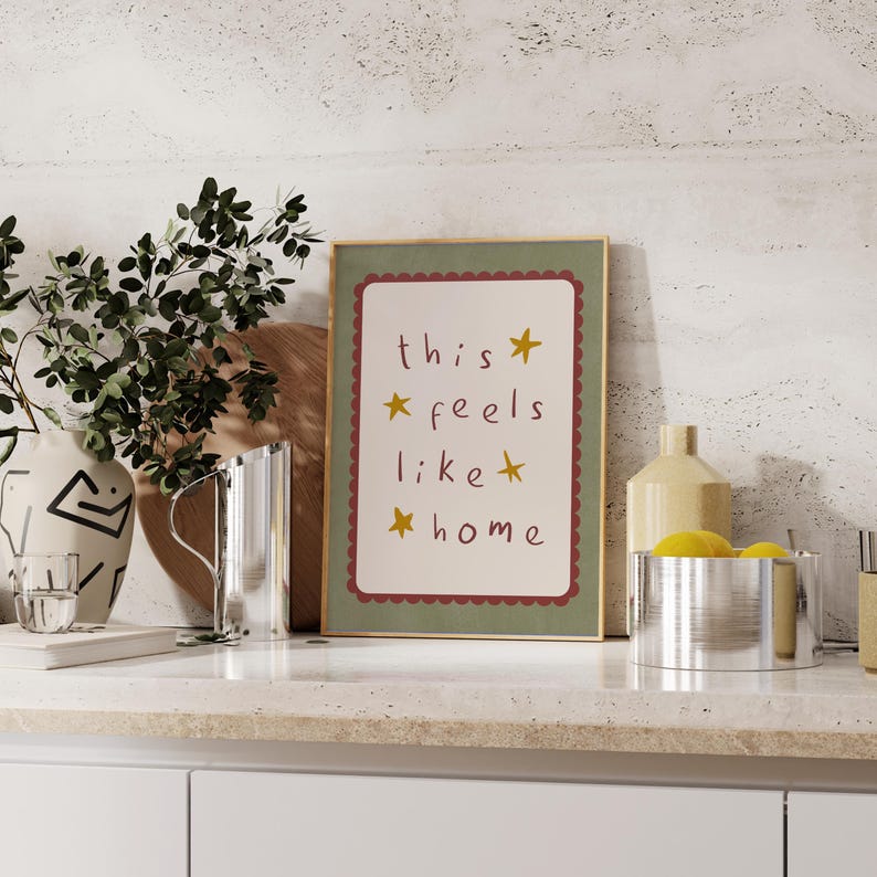 K&ouml;nnte beinhalten: Ein gerahmter Druck mit gr&uuml;nem Rahmen und wei&szlig;em Hintergrund. Der Druck zeigt den Text "this feels like home" in roter Handschrift mit gelben Sternen.