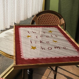 K&ouml;nnte beinhalten: Ein gerahmtes Schild mit einem rosa-wei&szlig;en gewellten Rand und den Worten "this feels like home" in Schwarz mit goldenen Sternen.
