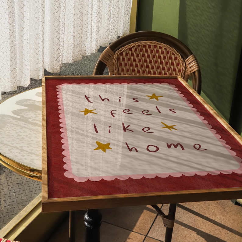 K&ouml;nnte beinhalten: Ein gerahmtes Schild mit einem rosa-wei&szlig;en gewellten Rand und den Worten "this feels like home" in Schwarz mit goldenen Sternen.