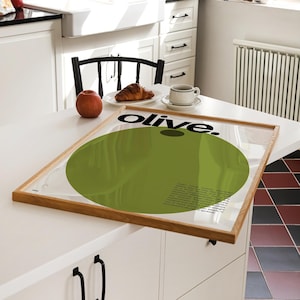 May include: A framed poster featuring a green circle and the word "olive." in black text. The poster is displayed on a white table with a red apple, a croissant, and a coffee cup.
