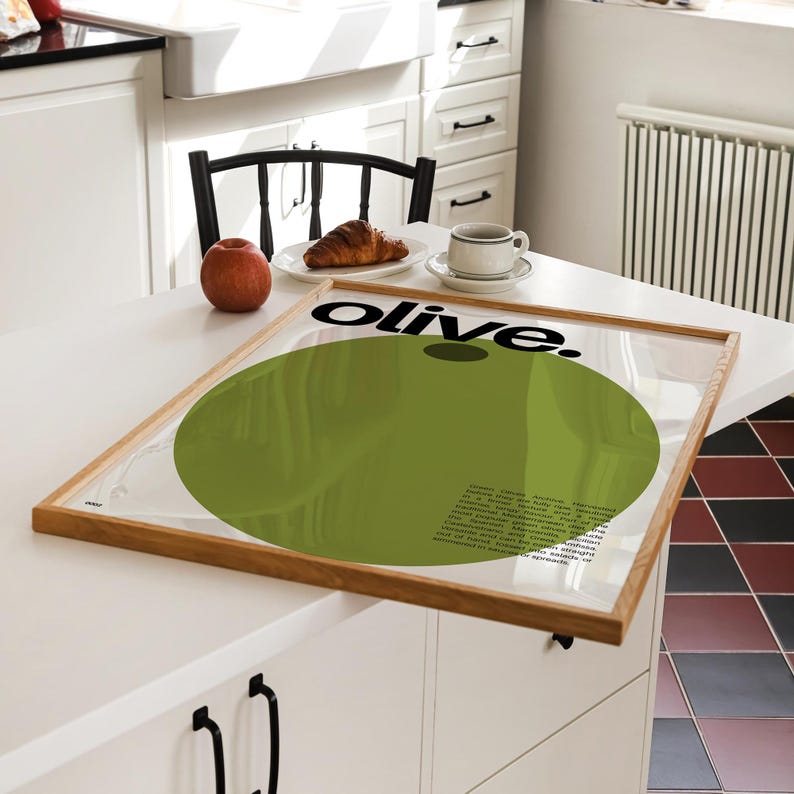 May include: A framed poster featuring a green circle and the word "olive." in black text. The poster is displayed on a white table with a red apple, a croissant, and a coffee cup.