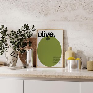 May include: A framed print featuring a green olive illustration and the word "olive." in black text. The print is displayed on a white countertop alongside a silver jug, a white vase with a black geometric design, and a silver bowl containing yellow lemons.
