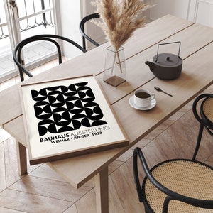 May include: A framed black and white geometric poster with the text "BAUHAUS AUSSTELLUNG WEIMAR - JUL-SEP. 1923" on a wooden table with a vase of dried pampas grass and a teapot.
