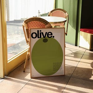 May include: A framed poster with the word "olive." in black text on a white background. A large green circle with a black dot in the centre is behind the text. The poster is leaning against a wooden chair in a cafe setting.