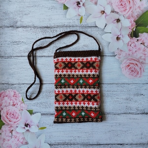 Puede incluir: Un bolso bandolera de punto con una correa marrón y un patrón geométrico en rojo, blanco, verde y marrón. El bolso es rectangular y tiene una parte superior marrón. El fondo presenta un arreglo floral.