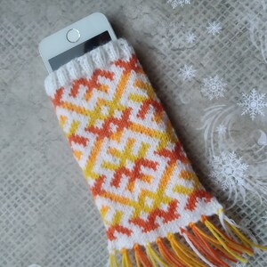 May include: A white smartphone in a knitted phone sleeve. The sleeve has a geometric pattern in orange and yellow on a white background, with fringe at the bottom. The sleeve is designed to protect the phone. The measurements are in centimetres.