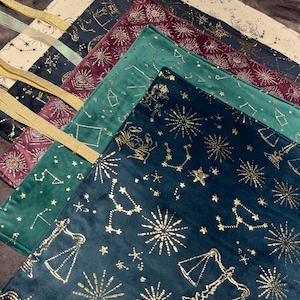 May include: A collection of velvet tarot cloths in various colors, including cream, navy, burgundy, and teal. Each cloth features a gold constellation design and a gold ribbon. The cloths are arranged in a layered display.