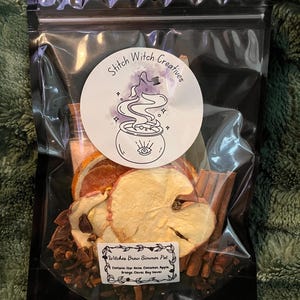 May include: A clear plastic bag filled with dried fruit and spices. The bag contains apple slices, cinnamon sticks, cloves, and orange slices. A round sticker with the text "Stitch Witch Creatives" and "Witches Brew Simmer Pot" is on the bag.