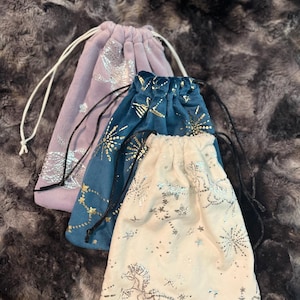 May include: Three drawstring bags in lavender, teal, and cream. Each bag features a silver celestial design with stars and a unicorn. The bags have white or black drawstrings and are made of a soft, velvety material.