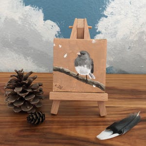 May include: A miniature painting of a bird perched on a branch, displayed on a small wooden easel. The artwork features a gray and white bird against a brown background. A pine cone and a feather are placed nearby.
