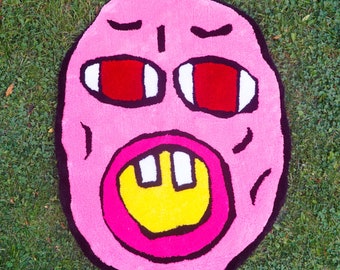 May include: A tufted rug shaped like an irregular oval face with a bright pink base. It features two large red eyes with white accents, a thick black outline, and a wide open mouth with a yellow interior and two prominent white rectangular teeth.