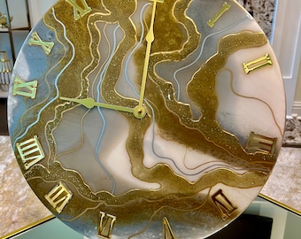 May include: A round, white and gold clock with Roman numeral markings. The clock face has a swirling, abstract design with gold and white accents. The clock is on a gold stand.