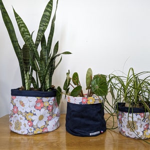 Könnte beinhalten: Drei Stoff-Pflanztöpfe mit Pflanzen. Die Töpfe sind marineblau mit einem Blumenmuster in Weiß, Gelb und Pfirsich. Die Pflanzen umfassen eine Bogenhanf, eine Peperomia und eine Ponytail-Palme. Die Töpfe stehen auf einem Holztisch.