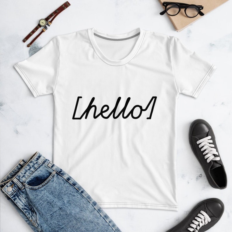 May include: White t-shirt with black text that says "[hello]" in a handwritten style. The shirt is laid flat on a white surface with a pair of jeans and a watch in the background.