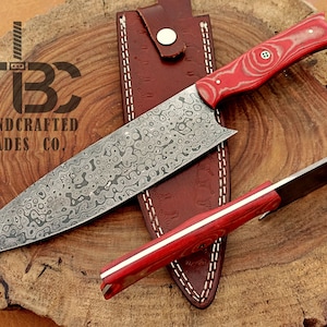 Puede incluir: Un cuchillo de chef hecho a mano con mango de madera roja y hoja de acero damasco. El cuchillo está sobre una tabla de cortar de madera con una funda de cuero.