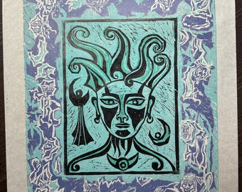 May include: A linocut print featuring a stylized face with teal and black details. The central image is framed by a border of teal and blue figures. The artwork is mounted on a white paper.