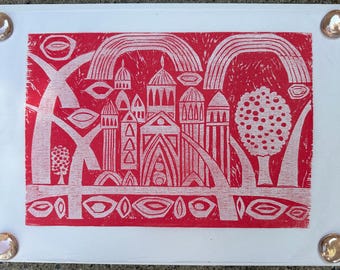 May include: Red and white linocut print featuring a stylized cityscape with arches, domes, and trees. The print has a folk art style, with a repeating pattern of shapes and lines. The print is mounted on white paper and held in place with four clear, round push pins.