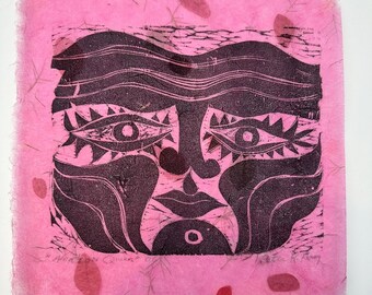 May include: A black and pink linocut print of a stylized face with large eyes, a nose, and a mouth. The print is on pink paper with a textured surface and small red accents. The text "African Queen" is visible.