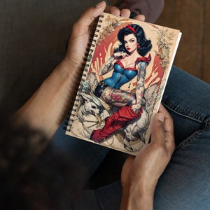 May include: A spiral bound notebook with a colorful illustration of a woman with tattoos and a wolf. The woman is wearing a blue and white corset and red boots. The background is a vintage style illustration.