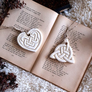 Könnte beinhalten: Zwei weiße Keramik-Anhänger in Herzform mit einem keltischen Knotendesign. Die Anhänger hängen an einer Schnur an einem Buch mit deutschem Text.
