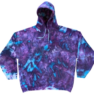 Puede incluir: Una sudadera con capucha y manga larga con un patrón de teñido anudado en tonos morado intenso, violeta oscuro y azul cian brillante. La prenda incluye un bolsillo tipo canguro frontal, capucha con cordones azul claro y puños acanalados.