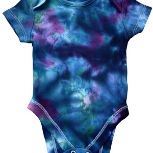 May include: A short-sleeved baby bodysuit featuring a tie-dye pattern in shades of deep blue, purple, and hints of green. The garment has a round neckline, white stitching along the seams, and two metal snap closures at the bottom crotch area.