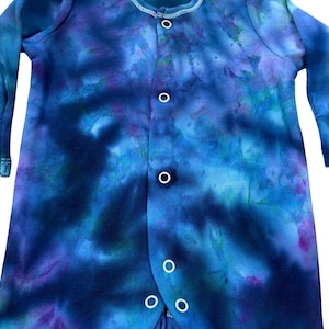 May include: A long-sleeved one-piece garment featuring a tie-dye pattern in shades of deep blue, purple, and teal. The front of the garment includes a vertical row of white snap fasteners extending from the round neckline down to the leg openings.