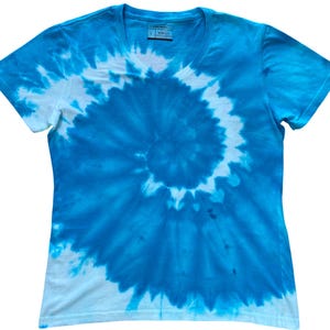 Könnte beinhalten: Ein kurzärmeliges T-Shirt mit V-Ausschnitt und einem blau-weißen Batik-Spiralmuster. Das Shirt zeigt ein zentrales Wirbeldesign, das von Dunkelblau zu helleren Tönen und Weiß übergeht. Ein Etikett mit der Größe L ist am Innenkragen sichtbar.