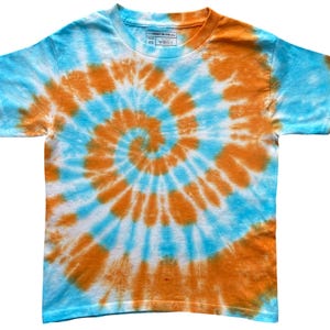 Op de afbeelding: Een t-shirt met korte mouwen en een ronde hals, voorzien van een levendig tie-dye spiraalpatroon in feloranje en hemelsblauw. Het kledingstuk heeft een standaard pasvorm met een ronde halslijn en een label aan de binnenzijde in maat XS.