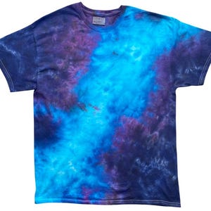 Puede incluir: Una camiseta de manga corta con cuello redondo y un patrón de teñido anudado en tonos morado intenso, azul marino oscuro y azul eléctrico brillante. La tela muestra una mezcla de colores arremolinados que van desde los bordes oscuros al centro.