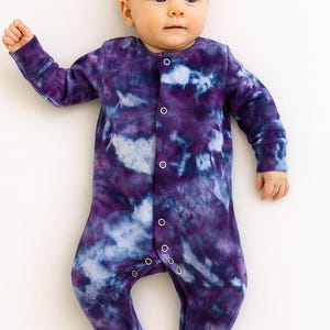Könnte beinhalten: Ein Baby in einem lila und blauen Tie-Dye-Fußstrampler. Das langärmlige Kleidungsstück hat eine Knopfleiste vorne und bedeckt die Füße des Babys. Das Baby liegt auf einer weißen Oberfläche.