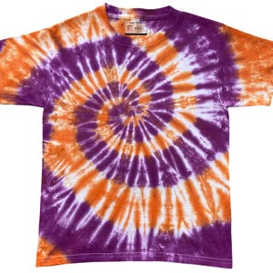 Könnte beinhalten: Ein kurzärmeliges T-Shirt mit Rundhalsausschnitt und einem lebendigen Tie-Dye-Spiralmuster in den Farben Violett, Orange und Weiß. Das Kleidungsstück hat eine Standardpassform mit einem sichtbaren Innenetikett, das den Text "YOUTH S" und "WASH" enthält.