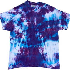 Puede incluir: Una camiseta de manga corta con un patrón tie-dye en tonos morado intenso, azul brillante y blanco. La prenda tiene un cuello redondo y un corte estándar, con el tinte creando un diseño abstracto y arremolinado sobre la tela.