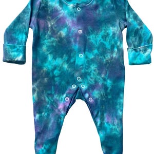 Op de afbeelding: Een romper met lange mouwen en voetjes, voorzien van een tie-dye patroon in tinten teal, donkerblauw en paars. Het kledingstuk heeft een ronde halslijn, lange mouwen met boorden en een verticale sluiting met witte drukknopen tot aan de voeten.
