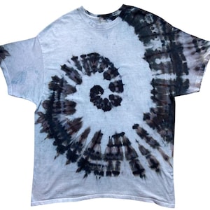 Op de afbeelding: Een t-shirt met korte mouwen en een ronde hals met een zwart en grijs tie-dye spiraalpatroon. Het ontwerp straalt vanuit het midden van het shirt en verloopt van een donkere zwarte kern naar lichtere grijze en witte tinten op de stof.