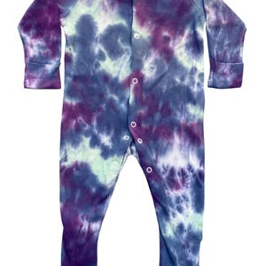 Peut inclure: Une grenouillère à manches longues avec un motif tie-dye dans des tons de violet profond, bleu marine et vert menthe pâle. Le vêtement présente une rangée verticale de boutons-pression blancs sur le devant, du col jusqu'à l'entrejambe.