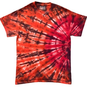 Op de afbeelding: Een t-shirt met korte mouwen voorzien van een levendig tie-dye patroon in tinten felrood, dieporanje en donkerzwart. Het ontwerp straalt vanaf de rechterkant uit in een zonnestraaleffect met duidelijke donkere strepen over de stof.