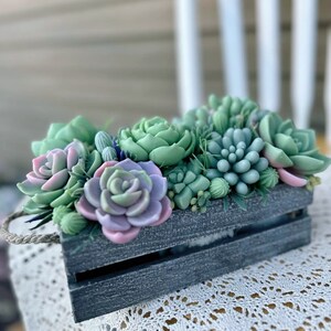 Puede incluir: Una peque&ntilde;a caja de madera llena de plantas suculentas artificiales. Las suculentas son de color verde, rosa y morado. La caja est&aacute; pintada de gris oscuro y tiene un asa de cuerda.