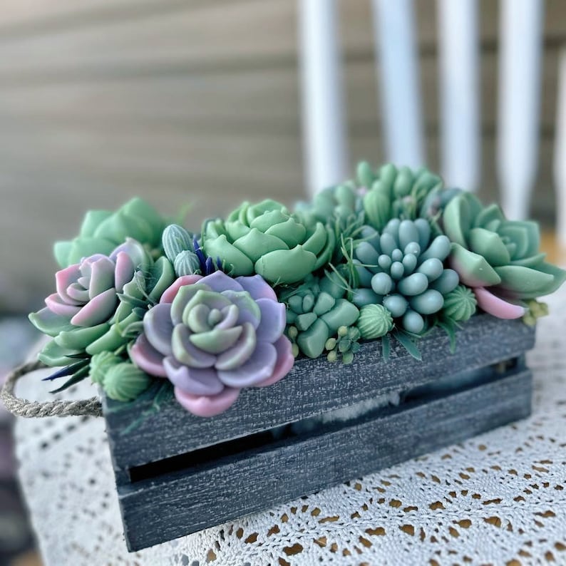 Puede incluir: Una peque&ntilde;a caja de madera llena de plantas suculentas artificiales. Las suculentas son de color verde, rosa y morado. La caja est&aacute; pintada de gris oscuro y tiene un asa de cuerda.