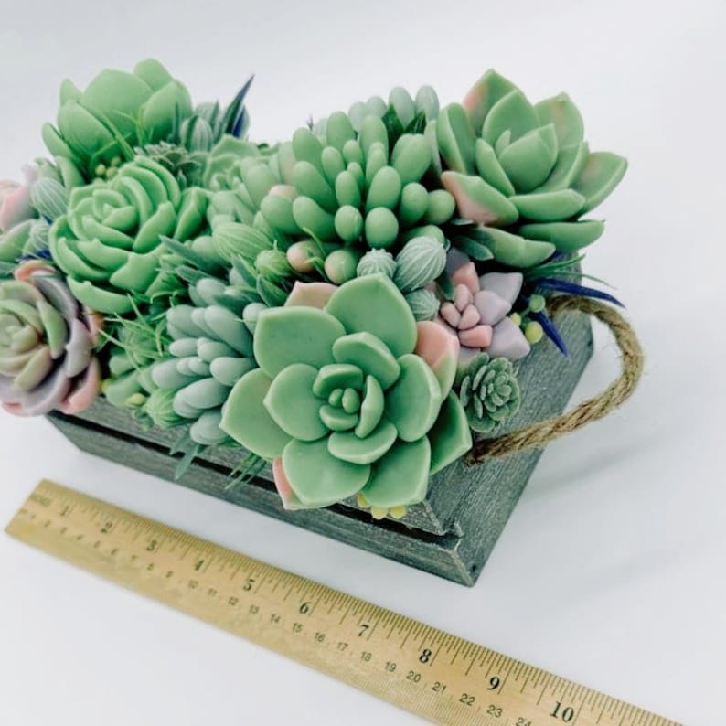 Puede incluir: Una peque&ntilde;a caja de madera con asa de cuerda llena de suculentas falsas en tonos verdes y rosas. La caja mide aproximadamente 20 cm de largo.