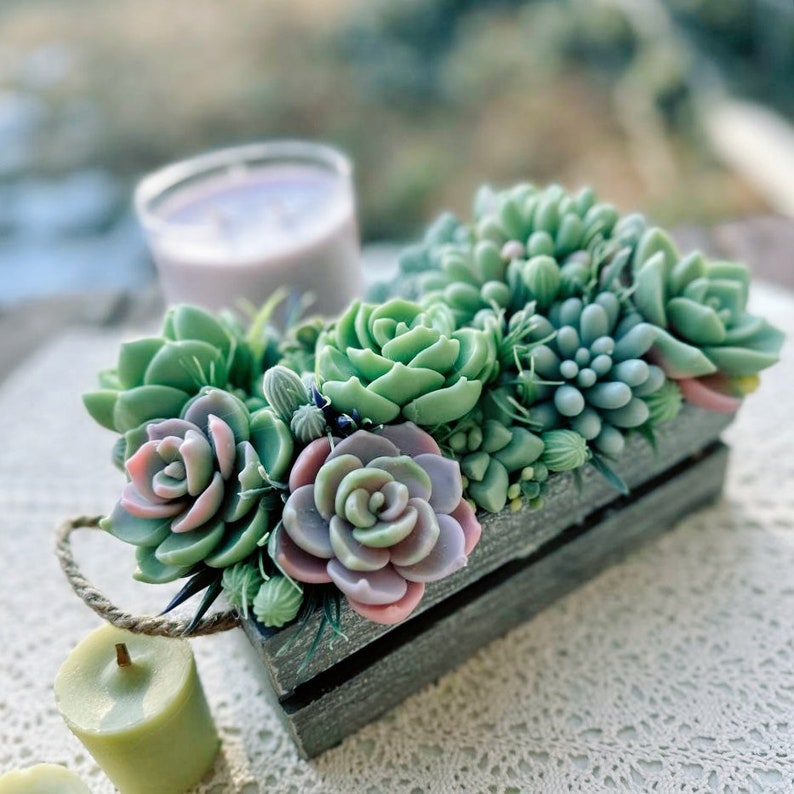 Puede incluir: Una caja de madera gris llena de jabones con forma de suculentas verdes y rosas. La caja tiene un asa de cuerda.