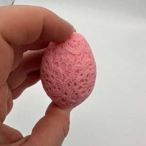 Puede incluir: Un objeto decorativo rosa en forma de huevo con un diseño intrincado y calado. La superficie presenta un patrón detallado de formas arremolinadas y motivos florales. El artículo se sostiene en una mano sobre un fondo blanco. La estética general es delicada y ornamentada.